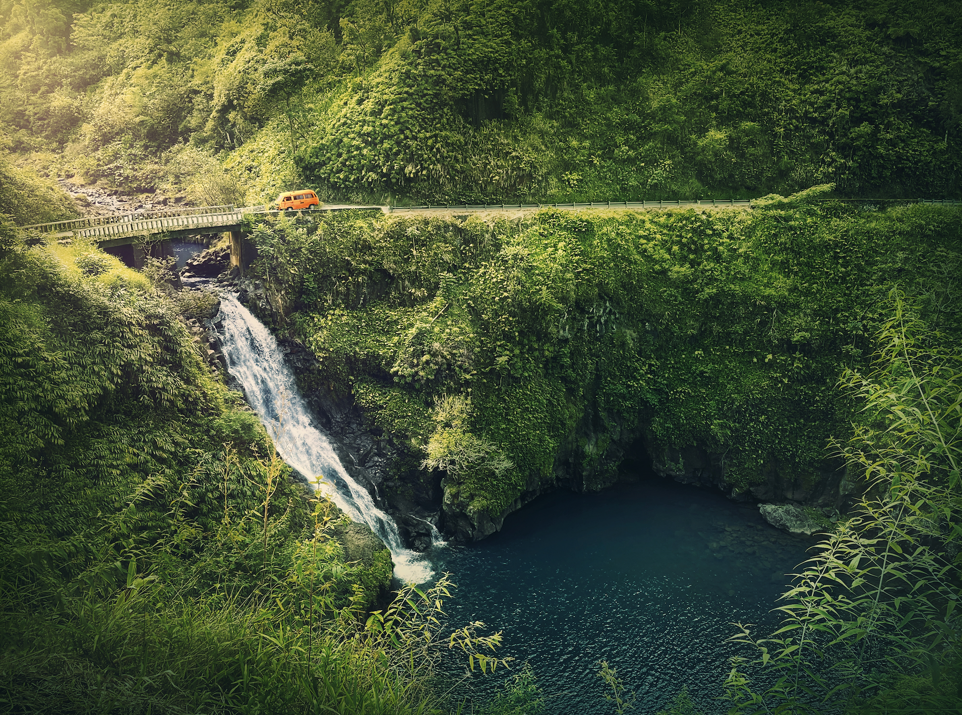 view of Makapipi Falls. waterfall under bridge as orange van drives along road 